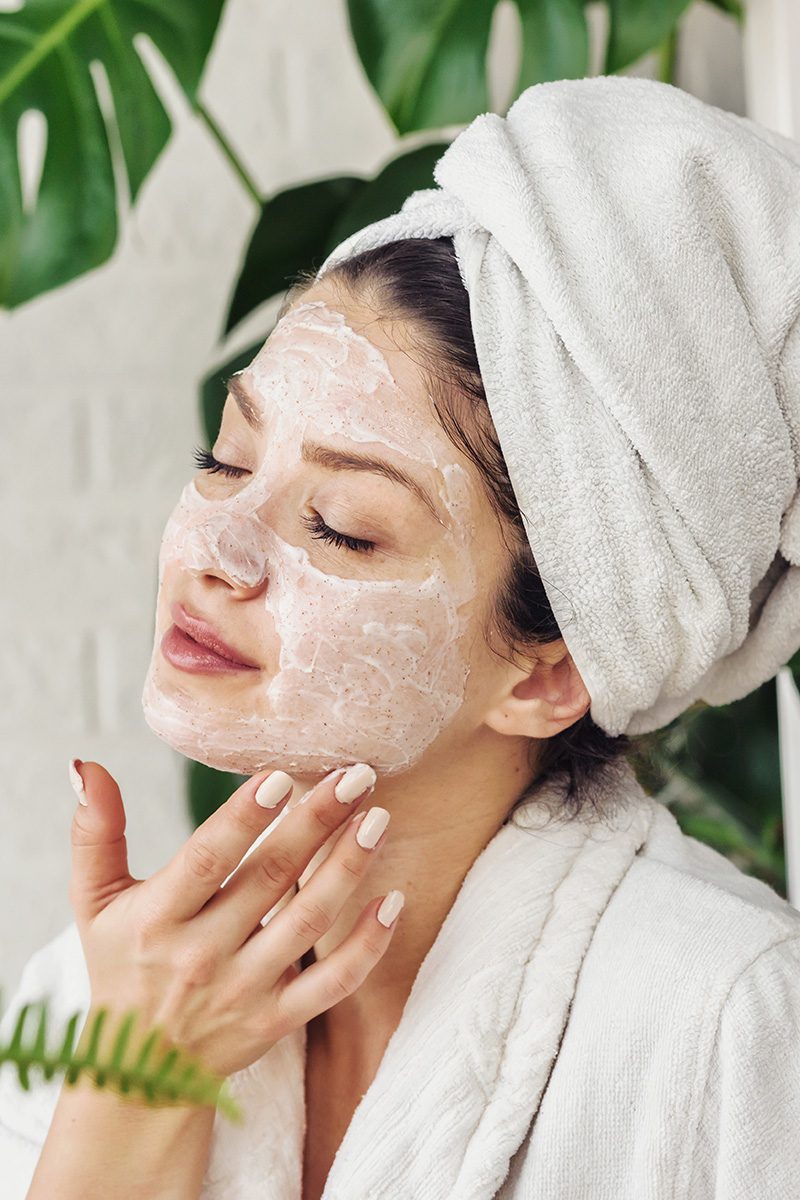 Frau vor Zimmerpflanze Monstera mit Bademantel, Handtuchturban und Gesichtsmaske, berührt ihr Gesicht